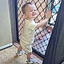 toddler, child, smiling, standing, indoor, door, mesh_door, brick_wall, carpet, shorts, shirt, footwear, checkered_shoes, hand_on_door, curtain, toy, happy, young_child, person, home