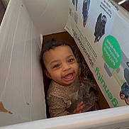Aliya is registered to the contest to win money with this photo: baby, brown_skin, cardboard_box, carpet, child, curly_hair, cute, eyes, face, happy, home, indoor, mouth, packaging, photograph, playful, portrait, sitting, smiling, sweater
