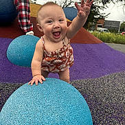 Sequoia is registered to the contest to win money with this photo: baby, child, playground, blue_sphere, textured_surface, happy, smiling, outdoor, person, trees, building, romper, hand, grass, colorful, checkered_pants, overcast_sky, reaching, fun, play