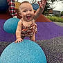 baby, child, playground, blue_sphere, textured_surface, happy, smiling, outdoor, person, trees, building, romper, hand, grass, colorful, checkered_pants, overcast_sky, reaching, fun, play