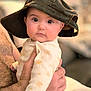 baby, child, hat, face, eyes, hand, adult_hand, indoor, portrait, clothing, infant, person, skin, cute, expression, warm_light, soft_focus, cozy, newborn, sitting