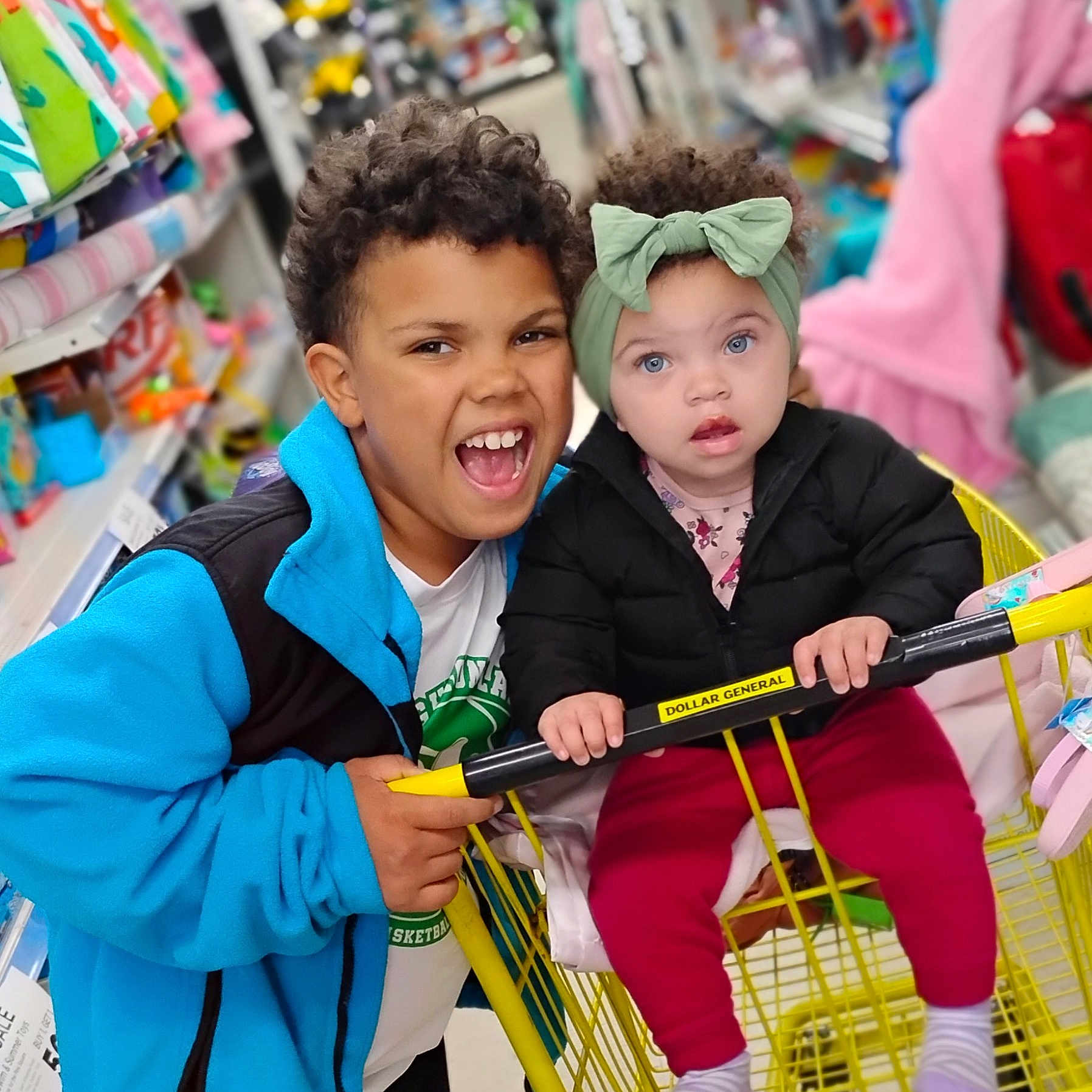 Keziah joined the competition — help win amazing prizes! aisle, baby, child, clothing, curly_hair, family, happy, headband, indoor, jacket, pants, person, retail, shopping_cart, sibling, smile, socks, store, toddler, yellow_cart