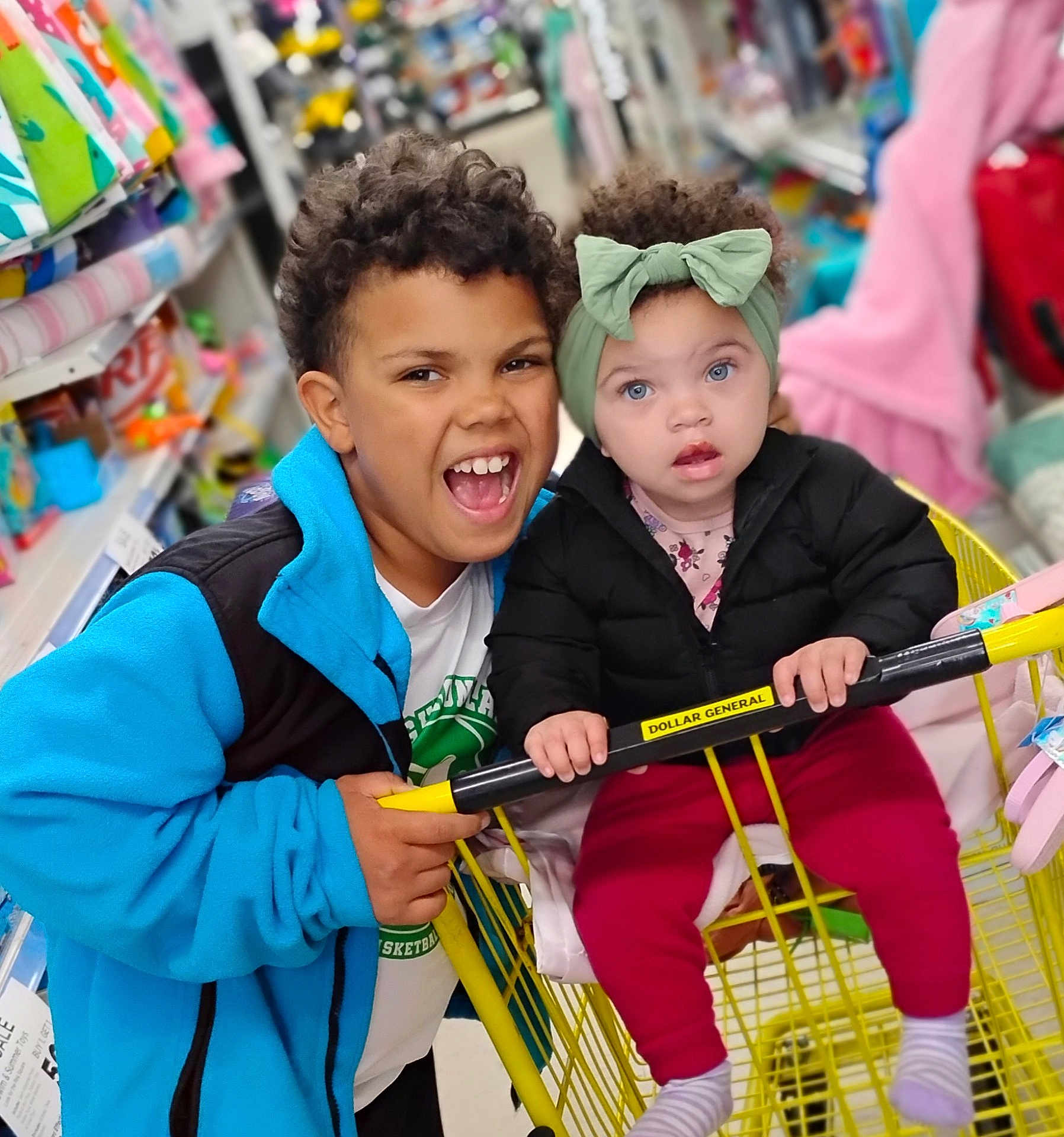 Keziah joined the competition — help win amazing prizes! child, baby, shopping_cart, store, aisle, clothing, headband, curly_hair, smile, happy, retail, indoor, yellow_cart, jacket, pants, socks, person, toddler, sibling, family