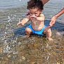 Keziah joined the competition — help win amazing prizes! toddler, child, water, splashing, beach, adult_hands, blue_swimwear, pebbles, outdoor, summer, playful, happy, nature, sunlight, water_droplets, barefoot, curly_hair, shallow_water, person, fun