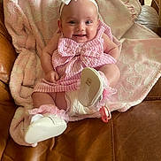 Alora is registered to the contest to win money with this photo: baby, infant, smiling, pink_dress, bow, headband, white_shoes, blanket, heart_pattern, leather_couch, happy, cute, indoors, child, pacifier, soft_texture, sitting, feet, person, cozy