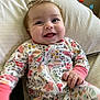 baby, infant, smiling, happy, onesie, floral_pattern, pillow, indoor, child, cute, face, skin, hand, person, baby_clothes, young, lying_down, cozy, expression, joy
