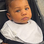 Khalil is registered to the contest to win money with this photo: baby, child, stroller, white_bib, curious_eyes, serious_expression, indoor, seatbelt, portrait, infant, clothing, person, face, head, skin, hand, furniture, black_stroller, closeup, young