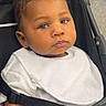 baby, child, stroller, white_bib, curious_eyes, serious_expression, indoor, seatbelt, portrait, infant, clothing, person, face, head, skin, hand, furniture, black_stroller, closeup, young
