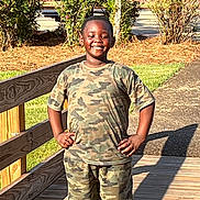 Kaiden is registered to the contest to win money with this photo: child, smiling, outdoor, portrait, standing, wooden_bridge, camouflage_clothing, shorts, sunlight, garden, plants, flowers, fence, pathway, shadow, daytime, happy, hands_on_hips, park, photography