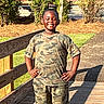 child, smiling, outdoor, portrait, standing, wooden_bridge, camouflage_clothing, shorts, sunlight, garden, plants, flowers, fence, pathway, shadow, daytime, happy, hands_on_hips, park, photography