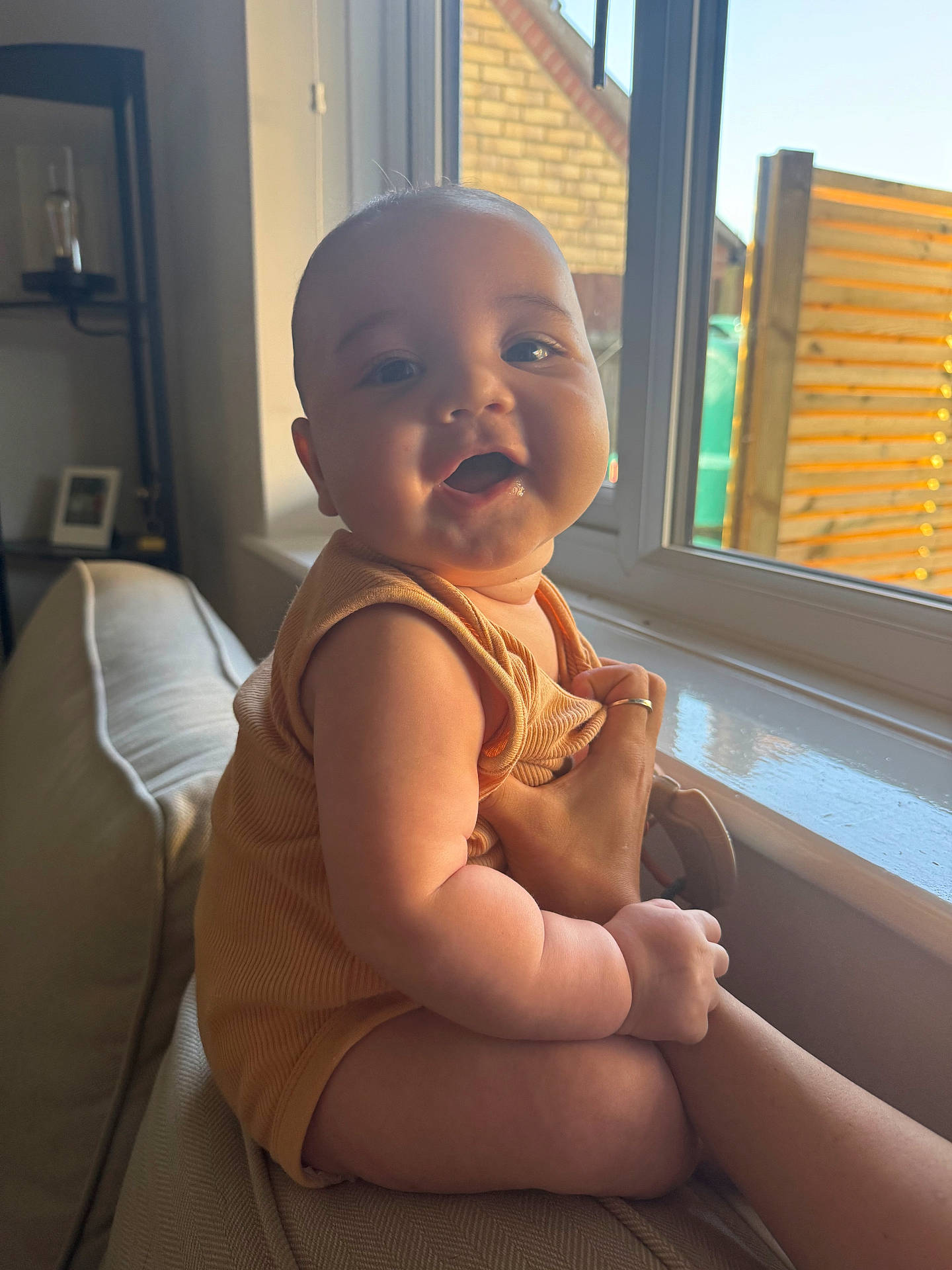arm, baby, child, clothing, couch, cozy, face, furniture, hand, home, indoor, infant, person, sitting, skin, smiling, sunlight, warm_light, window, window_frame