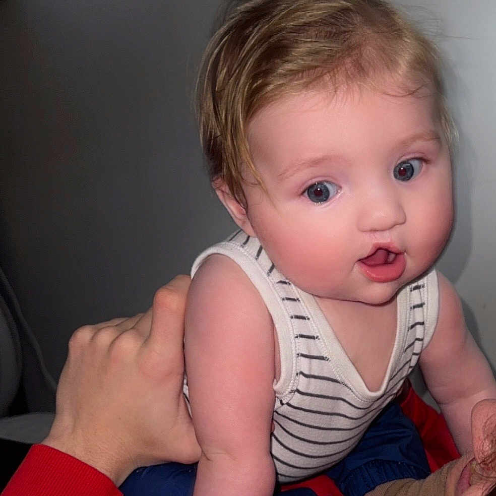 Astin joined the competition — help win amazing prizes! baby, blue_eyes, child, close_up, clothing, curly_hair, cute, expression, face, hand, holding, human, indoor, infant, person, portrait, red_sweatshirt, skin, striped_tank_top, young