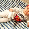 Nolan a rejoint le concours — aidez-le/la à gagner de superbes lots ! baby, child, superman, red_hat, white_clothing, striped_rug, floor_tiles, indoor, lying_down, cute, portrait, infant, young_child, smiling, person, headwear, footwear, home, relaxed, pose