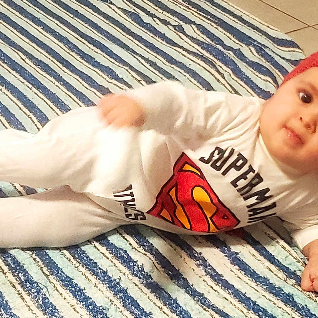 Nolan a rejoint le concours — aidez-le/la à gagner de superbes lots ! baby, child, superman, red_hat, white_clothing, striped_rug, floor_tiles, indoor, lying_down, cute, portrait, infant, young_child, smiling, person, headwear, footwear, home, relaxed, pose