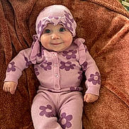 Oaklynn is registered to the contest to win money with this photo: baby, child, infant, smile, purple_clothing, flower_pattern, knitwear, blanket, soft_texture, cute, happy, portrait, indoors, headwear, cozy, lying_down, small_hands, buttoned_sweater, warm_colors, comfortable