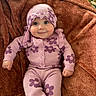 baby, child, infant, smile, purple_clothing, flower_pattern, knitwear, blanket, soft_texture, cute, happy, portrait, indoors, headwear, cozy, lying_down, small_hands, buttoned_sweater, warm_colors, comfortable