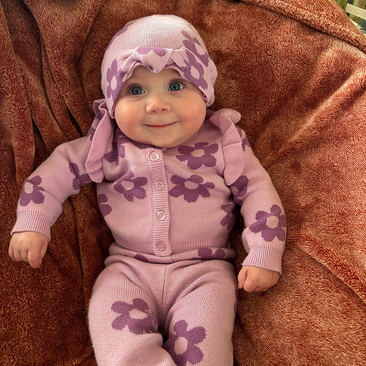 Oaklynn is registered to the contest to win money with this photo: baby, child, infant, smile, purple_clothing, flower_pattern, knitwear, blanket, soft_texture, cute, happy, portrait, indoors, headwear, cozy, lying_down, small_hands, buttoned_sweater, warm_colors, comfortable