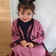 Joudia participe au concours pour gagner de l'argent avec cette photo : child, smile, hair_buns, pink_clothing, black_trim, embroidery, sitting, floor, wooden_floor, indoor, wall, cute, portrait, young_child, person, happy, casual, home, girl, fashion