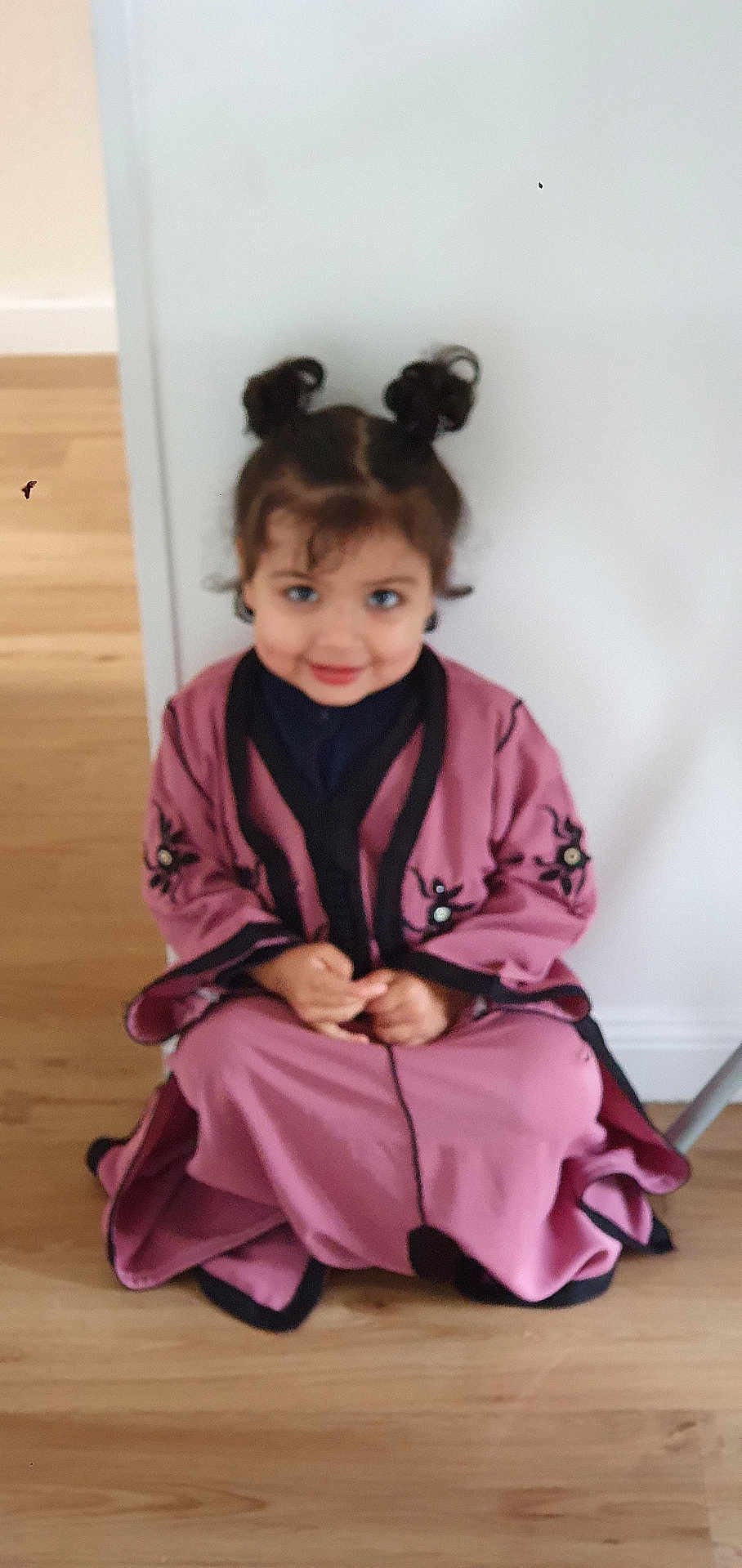 Joudia participe au concours pour gagner de l'argent avec cette photo : child, smile, hair_buns, pink_clothing, black_trim, embroidery, sitting, floor, wooden_floor, indoor, wall, cute, portrait, young_child, person, happy, casual, home, girl, fashion