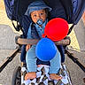 baby, infant, child, stroller, pacifier, sun_hat, balloons, red_balloon, blue_balloon, outdoor, portrait, sitting, baby_clothing, blue_clothing, blanket, sidewalk, pavement, wheel, sunlight, happy
