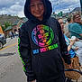 child, smiling, hoodie, colorful, outdoor, cloudy_sky, street, bench, woman, baby, bottle, feeding, people, casual_clothing, sidewalk, shopping_bag, trees, cars, buildings, urban