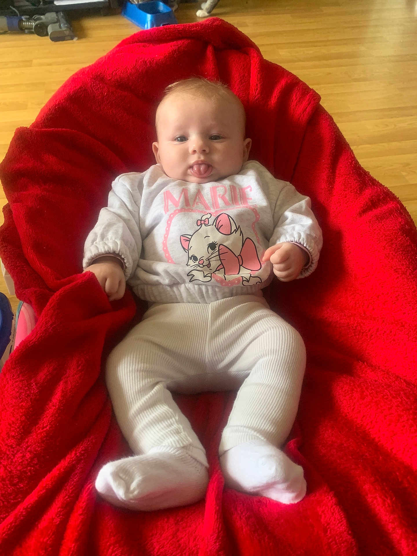 Inaya a rejoint le concours — aidez-le/la à gagner de superbes lots ! baby, infant, child, red_blanket, white_socks, cartoon_sweater, tongue_out, wooden_floor, cozy, cute, playful, indoor, smiling, sitting, person, face, clothing, plush, comfortable, young