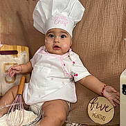 Melanie is registered to the contest to win money with this photo: baby, infant, chef_hat, apron, whisk, flour, baking, kitchen, food, bread, table, cute, child, sitting, face, expression, milk_bottle, wooden_sign, cloth_background, red_bracelet