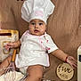 baby, infant, chef_hat, apron, whisk, flour, baking, kitchen, food, bread, table, cute, child, sitting, face, expression, milk_bottle, wooden_sign, cloth_background, red_bracelet
