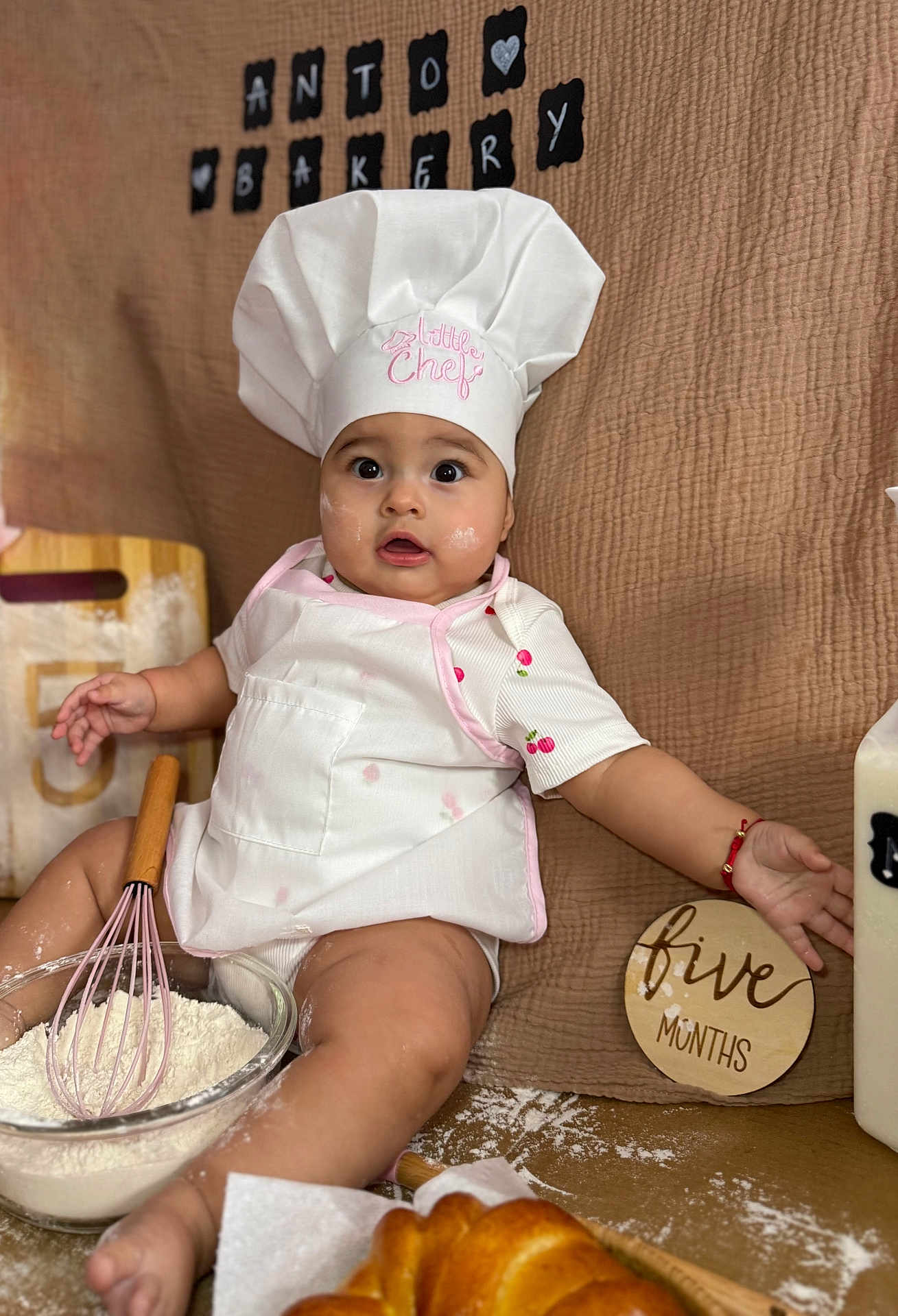 Melanie is registered to the contest to win money with this photo: baby, infant, chef_hat, apron, whisk, flour, baking, kitchen, food, bread, table, cute, child, sitting, face, expression, milk_bottle, wooden_sign, cloth_background, red_bracelet