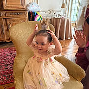 Sofia participe au concours pour gagner de l'argent avec cette photo : toddler, child, dress, crown, chair, indoor, carpet, wooden_floor, furniture, smile, person, playful, decor, table, lamp, window, curtain, hand, face, happy