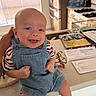 baby, smiling, child, kitchen, denim_overalls, striped_shirt, socks, toy, counter, papers, refrigerator, stove, hand, window_blinds, happy, indoor, person, face, infant, home