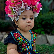 Emily joined the competition — help win amazing prizes! toddler, child, flower_crown, traditional_dress, embroidered_clothing, outdoor, portrait, greenery, nature, baby, cute, serious_expression, colorful, sitting, person, young_child, headwear, garden, young_kid, closeup