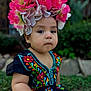 toddler, child, flower_crown, traditional_dress, embroidered_clothing, outdoor, portrait, greenery, nature, baby, cute, serious_expression, colorful, sitting, person, young_child, headwear, garden, young_kid, closeup