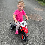Mya participe au concours pour gagner de l'argent avec cette photo : child, toddler, tricycle, toy_vehicle, pink_shirt, outdoor, road, asphalt, casual_clothing, short_sleeves, footwear, curly_hair, expression, sidewalk, greenery, manhole_cover, daylight, small_vehicle, playtime, recreation