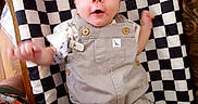 Remington is registered to the contest to win money with this photo: baby, child, infant, lying_down, portrait, face, wide_eyed, outfit, overalls, gray_clothing, checkerboard, mat, wooden_table, indoor, home, arms, legs, smiling, bottle, household_items