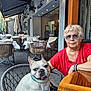 woman, dog, french_bulldog, outdoor, restaurant, table, chair, sunglasses, red_shirt, wicker_chair, pet, smiling_dog, casual, daylight, urban, pavement, glassware, umbrellas, relaxation, companion