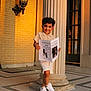 Santiago is registered to the contest to win money with this photo: child, boy, smiling, curly_hair, shorts, sneakers, newspaper, column, brick_wall, door, outdoor, evening_light, architecture, concrete_floor, pose, happy, casual_clothing, portrait, standing, urban