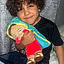 child, curly_hair, smile, plush_doll, tablet, black_shirt, blue_jeans, bed, curtain, indoor, seated, toy, happy, portrait, person, furniture, hand, cuddling, casual, home
