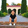 child, boy, sitting, stone_steps, black_shirt, denim_shorts, white_sneakers, garden, pathway, trees, archway, greenery, outdoor, daylight, smile, curly_hair, casual_clothing, nature, relaxed, portrait