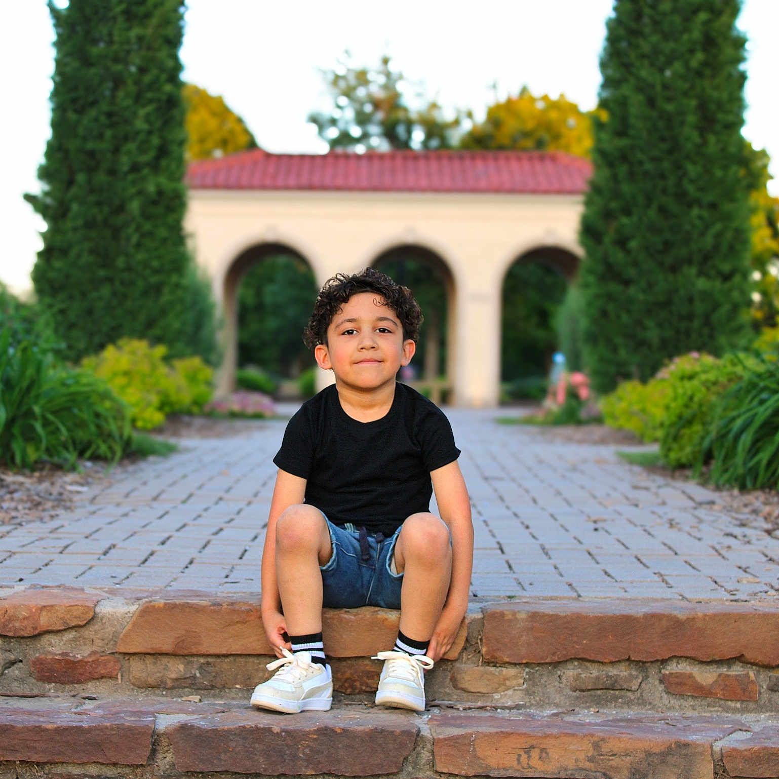 Santiago joined the competition — help win amazing prizes! archway, black_shirt, boy, casual_clothing, child, curly_hair, daylight, denim_shorts, garden, greenery, nature, outdoor, pathway, portrait, relaxed, sitting, smile, stone_steps, trees, white_sneakers