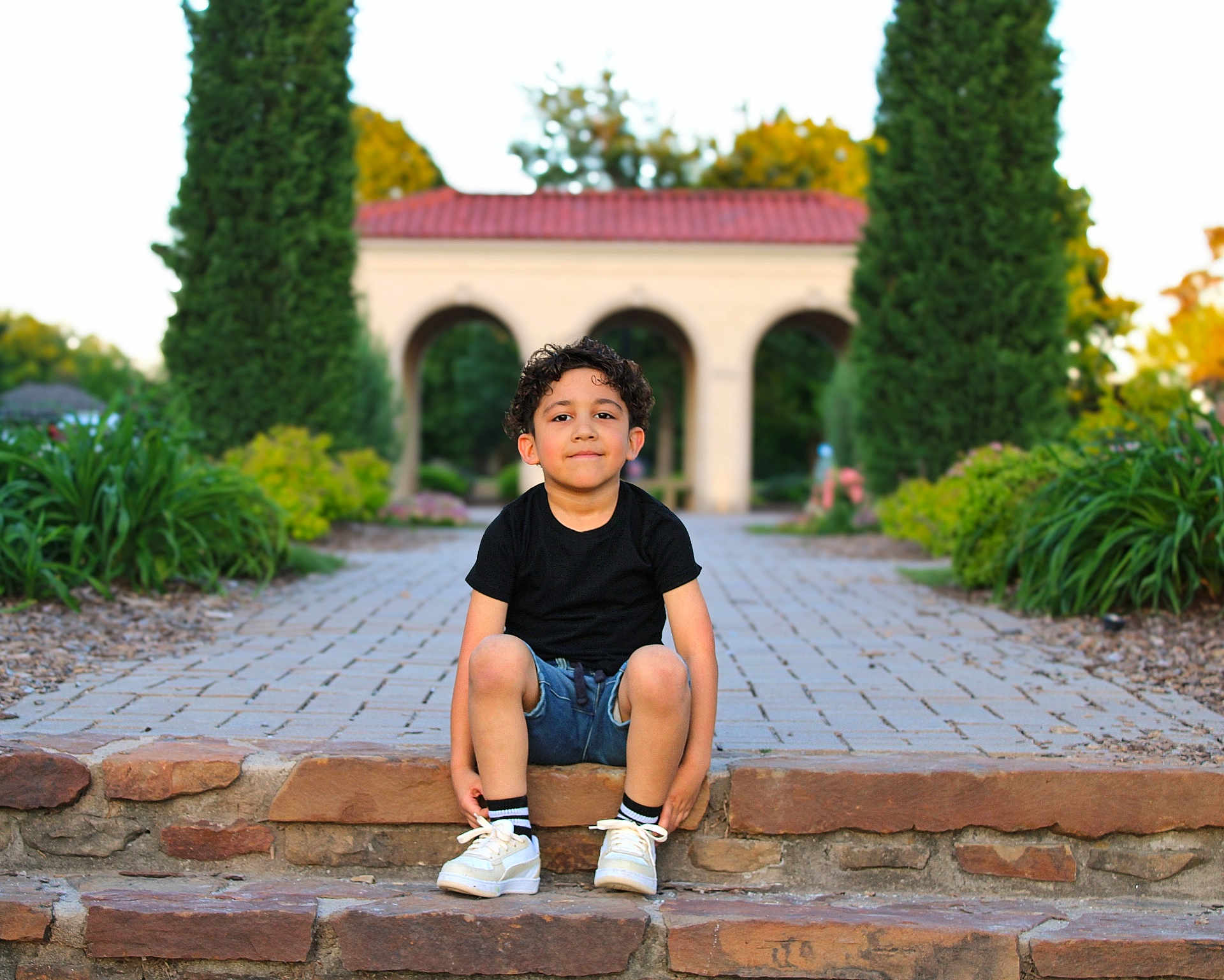 Santiago joined the competition — help win amazing prizes! child, boy, sitting, stone_steps, black_shirt, denim_shorts, white_sneakers, garden, pathway, trees, archway, greenery, outdoor, daylight, smile, curly_hair, casual_clothing, nature, relaxed, portrait