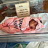 Jream is registered to the contest to win money with this photo: newborn, baby, hospital, bassinet, sleeping, pink_outfit, headband, bow, infant, blanket, sign, birth_details, crib, clothing, person, cute, resting, indoors, medical, portrait