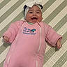 baby, child, smiling, happy, portrait, lying_down, pink_outfit, onesie, headband, bow, blanket, bed, indoor, infant, face, hands, clothing, patterned_background, sleepwear, cute
