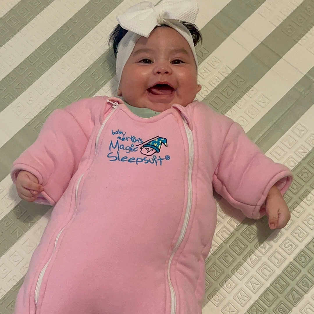 Dayamberly Garcia is registered to the contest to win money with this photo: baby, child, smiling, happy, portrait, lying_down, pink_outfit, onesie, headband, bow, blanket, bed, indoor, infant, face, hands, clothing, patterned_background, sleepwear, cute