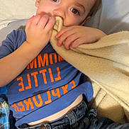 Asher is registered to the contest to win money with this photo: child, blanket, denim, blue_shirt, orange_text, face, hand, jeans, lying_down, curious_expression, soft_texture, comfort, casual_clothing, indoors, young_child, portrait, warm_lighting, relaxed, person, cozy