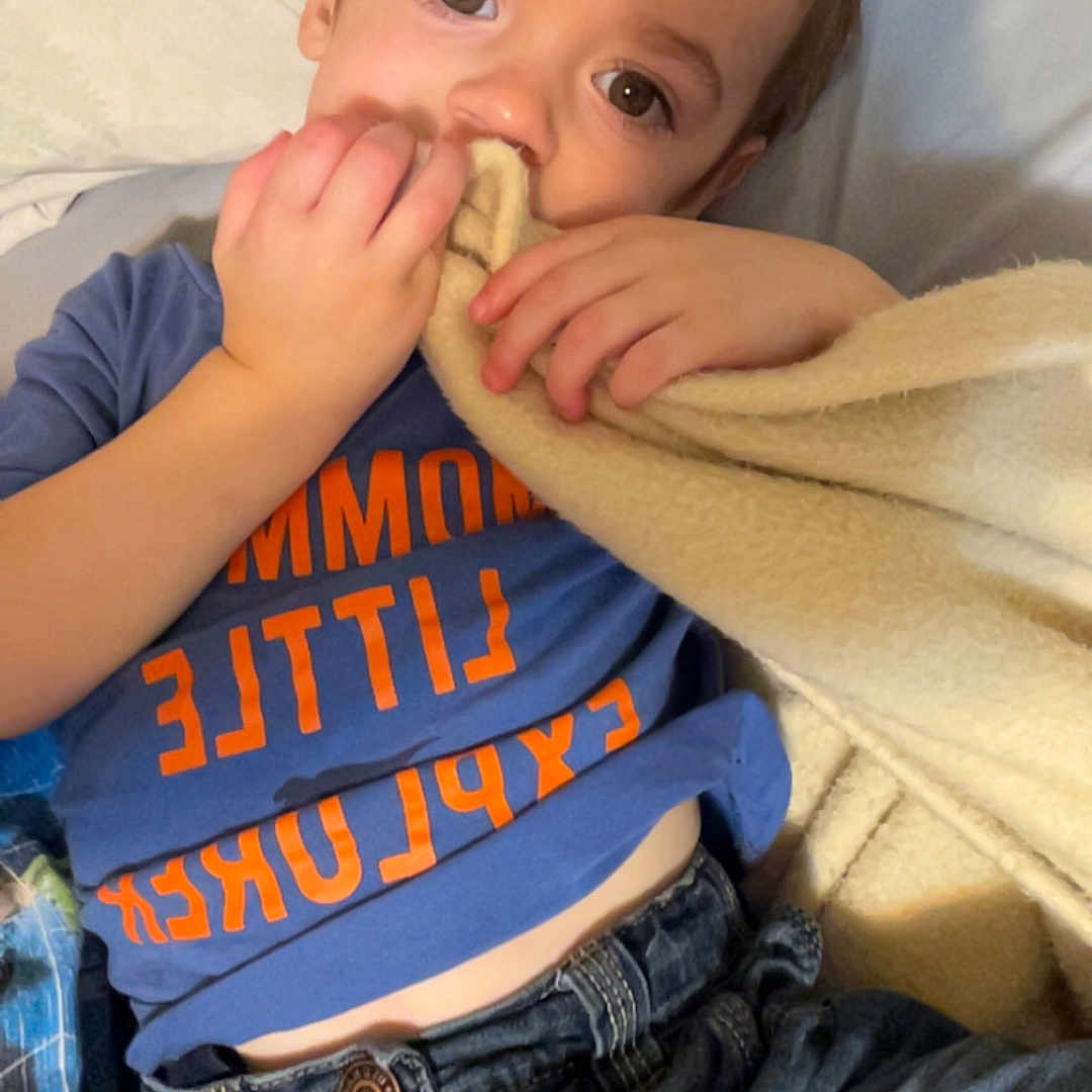 Asher is registered to the contest to win money with this photo: blanket, blue_shirt, casual_clothing, child, comfort, cozy, curious_expression, denim, face, hand, indoors, jeans, lying_down, orange_text, person, portrait, relaxed, soft_texture, warm_lighting, young_child