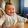 baby, smiling, high_chair, bottle, bib, table, indoor, child, cute, happy, feeding, wooden_furniture, person, face, infant, milk_bottle, casual_clothing, home, portrait, early_childhood