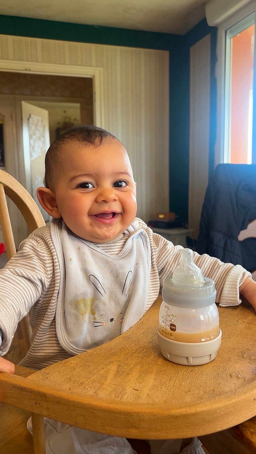 Tao participe au concours pour gagner de l'argent avec cette photo : baby, smiling, high_chair, bottle, bib, table, indoor, child, cute, happy, feeding, wooden_furniture, person, face, infant, milk_bottle, casual_clothing, home, portrait, early_childhood