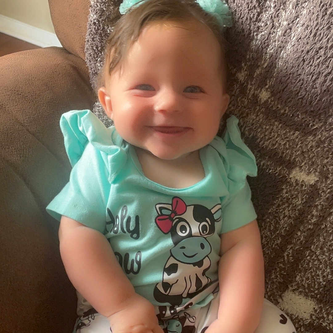 Faye is registered to the contest to win money with this photo: baby, blue_hair_accessories, cartoon_cow, child, clothing, comfort, cozy, cute, cute_outfit, furniture, hair_bows, happy, indoors, person, portrait, seat, sitting, smiling, soft_texture, young_child