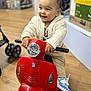 Mirán a rejoint le concours — aidez-le/la à gagner de superbes lots ! background, casual_clothing, child, cute, face, feet, flooring, handlebars, happy, head, indoor, person, play, red, shoes, smiling, store, sweater, toddler, toy_scooter